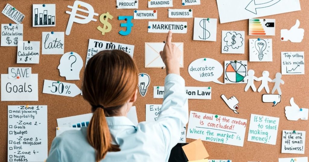 A women pointing towards the marketing writing on the wall. There are several marketing channels written in post-it notes.