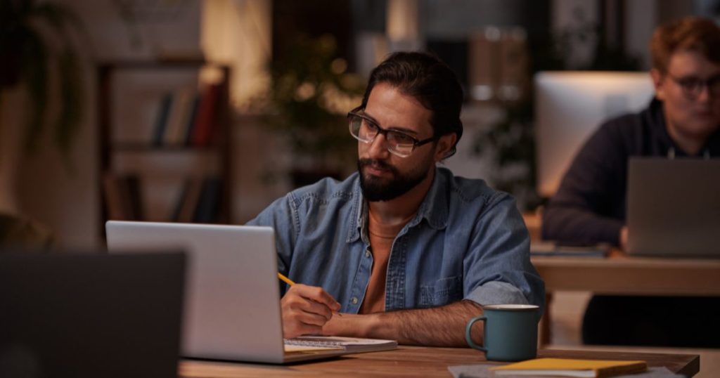 A man working on a laptop, while taking notes on a notebook.