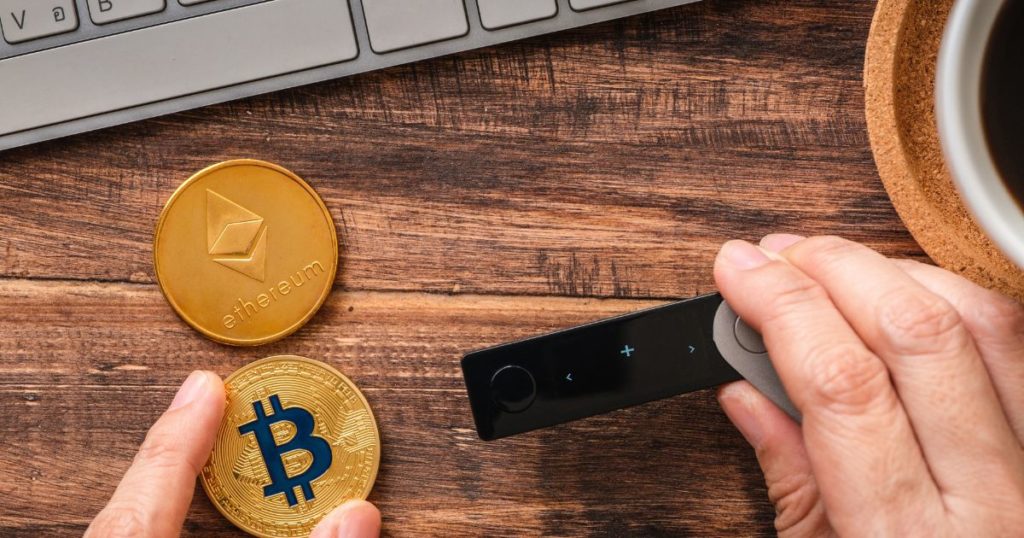 The right hand is holding a blockchain crypto wallet, while the left hand is holding a coin with the Bitcoin symbol on it. There is a physical coin with the Ethereum text and logo on the background. 